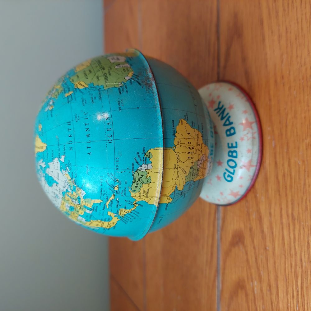 A vintage globe with a blue ocean map and yellow/green landmasses, displayed on a white round base printed with 'GLOBE BANK' and set on a wooden tabletop.